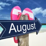 Wooden beach signpost reading August with pink flip-flops, summer sky