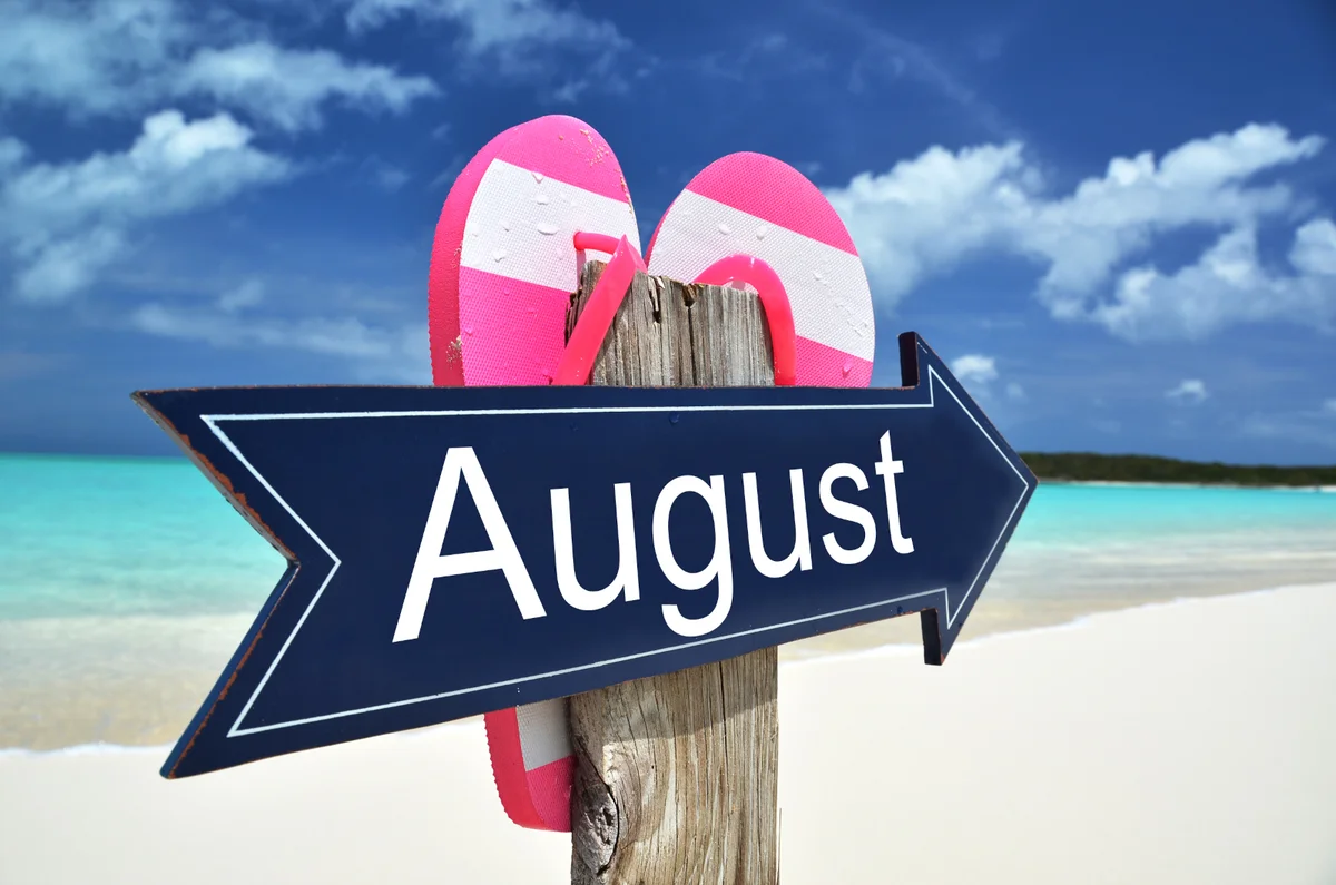 Wooden beach signpost reading August with pink flip-flops, summer sky